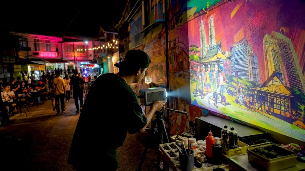 Seorang seniman sedang menyiapkan projek seni urban menggunakan teknik projection mapping di kawasan bandar.