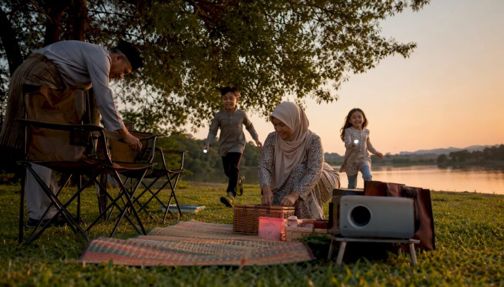 Sekeluarga sedang sibuk membuat persiapan untuk tayangan wayang terbuka di taman, lengkap dengan tikar dan makanan ringan untuk dinikmati bersama.